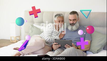 Liegendes reifes Paar, das sich ein Tablet auf dem Bett im Schlafzimmer teilt, Mann mit Bart, Frau in gestreiftem Oberteil Stockfoto