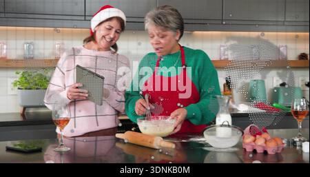 Frauen in festlicher Kleidung und Weihnachtsmannmütze, die auf der Kücheninsel mit Schneebesen und Tablette zum Rühren von Teig machen Stockfoto