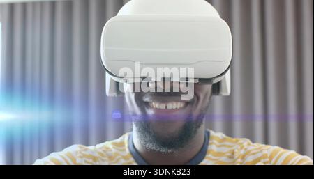 Lächelnder Mann mit weißem VR-Headset, das mit virtueller Szene zu Hause interagiert, gestreiftes T-Shirt Stockfoto