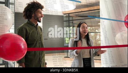 Sprechende Frau, lächelnder Mann in olivfarbenem Hemd grauem Mantel hinter rotem Band in Lobbyballons [GESCHWÄRZT] Stockfoto