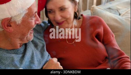 Lächelndes Seniorenpaar mit warmen Blicken zu Hause auf der Couch, mit Weihnachtsmütze, Pullover und Schmuck Stockfoto