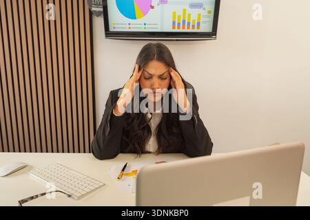 Junge Frau, die Schmerzen und Erschöpfung durch die Arbeit empfindet, überwältigende Kopfschmerzen an ihrem Schreibtisch Stockfoto