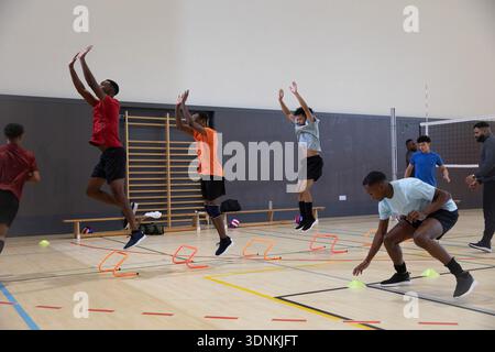 Afroamerikanische männliche Teenager-Athleten, die in der Turnhalle der Schule Agility Drilling über orange Mini Hürden durchführen Stockfoto