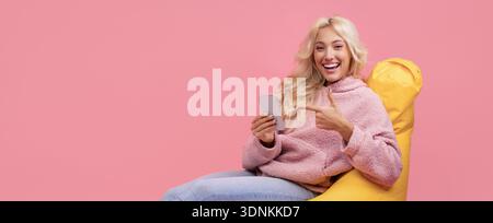Die junge Frau lächelt und zeigt auf ihr Telefon, während sie auf einem hellen gelben Stuhl sitzt Stockfoto