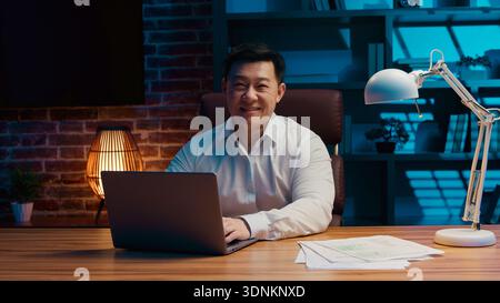 Fröhlicher ethnischer Mann mit Laptop im Abendbüro asiatischer glücklicher Geschäftsmann, der nachts mit Computer arbeitet, der in die Kamera lächelt koreanischer Geschäftsmann genießt Stockfoto