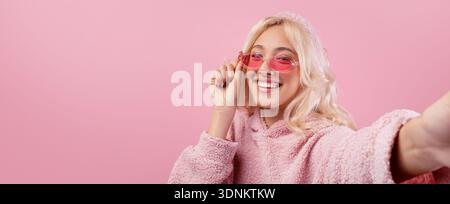 Die junge Frau macht tagsüber ein Selfie mit rosa Brille auf rosa Hintergrund Stockfoto