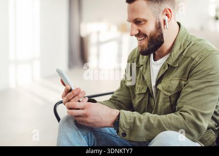 Seitenansicht Des Fröhlichen Kerl Mit Smartphone Tragen Ohrstöpsel In Innenräumen Stockfoto