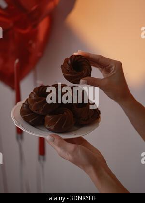 Nahaufnahme von weiblichen Händen, die einen weißen Teller mit köstlichen Schokoladenmuffins halten. Warme Lichtprojektion und rote Herzballons im Hintergrund für r Stockfoto