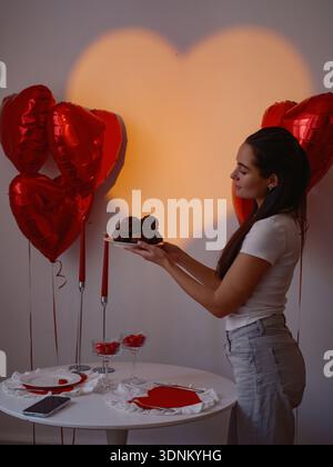Schöne junge Frau, die Teller mit Schokoladenmuffins in der Nähe eines romantischen Esstisch mit Herzservietten und roten Ballons unter warmem Herz hält Stockfoto