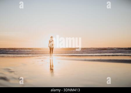 Frau, die bei Sonnenaufgang an einem sonnendurchfluteten Strand steht, mit Reflexion auf dem nassen Sand Stockfoto