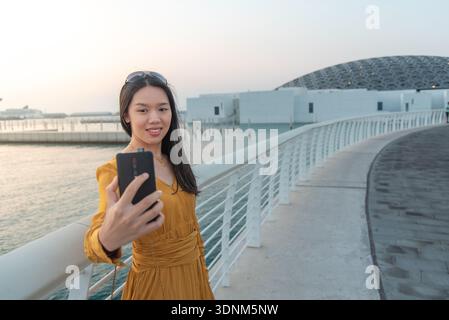 Eine modische junge asiatische Frau in gelbem Outfit macht ein Selfie vor dem Museumsbereich in Abu Dhabi in den Vereinigten Arabischen Emiraten mit der berühmten Kuppel A Stockfoto