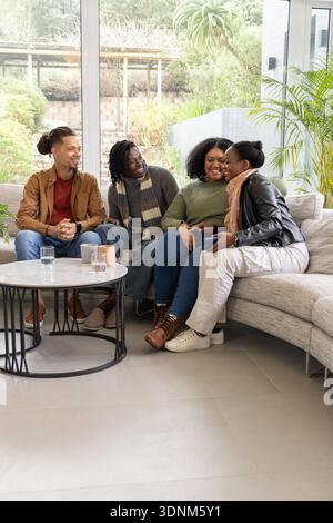 Verschiedene Freunde sitzen in der Nähe auf einem hellgrauen Sofa in der Nähe des Fensters und halten Smartphone und Tassen Stockfoto