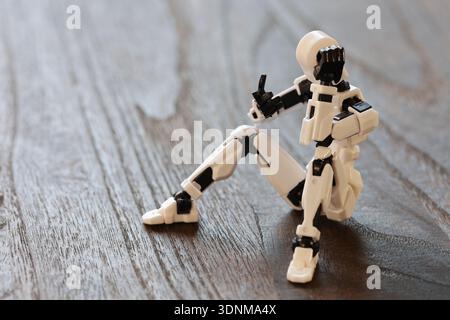 Eine kleine humanoide Figur sitzt lässig auf einer Holzoberfläche und zeigt eine nachdenkliche Facepalm-Pose, die einen unbeschwerten Blick auf futuristisches Design einfängt Stockfoto