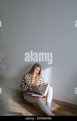 Frau, die mit einem Hardcover-Buch studiert, während sie auf dem Boden in der Nähe der weißen Wand sitzt. Minimales Innenraumkonzept, Lern- und Entspannungskonzept. Stockfoto