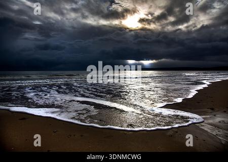 DRAMATISCHE ABENDLICHE AUFNAHME EINES DORSET UK STRANDES NACH EINEM STURM MIT DUNKLEN MOODY WOLKEN UND WEISSEM SCHAUM IN DEN MEERESWELLEN. Stockfoto