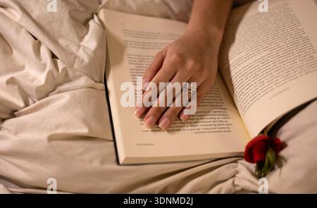 Frauenhand mit Verlobungsring auf einem offenen Buch im Bett, mit einer roten Rose Stockfoto