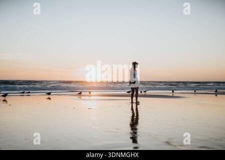 Frau, die an einem ruhigen Strand bei Sonnenuntergang mit Wellen, Möwen und Reflexionen steht Stockfoto
