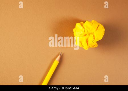 Gelbe zerknitterte Papierkugel und gelber Bleistift auf braunem Hintergrund. Stockfoto