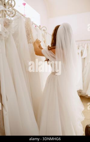 Eine junge Frau wählt ein Kleid in einem Brautsalon. Sie trägt ein Hochzeitskleid und einen Schleier und sieht sich andere Hochzeitskleider an. Eine Braut wählt ein Kleid Stockfoto