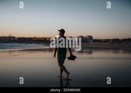 Mann, der bei Sonnenuntergang am Strand entlang geht, hält seinen Hut mit der Skyline der Stadt im Hintergrund Stockfoto