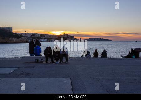 Marseille, Frankreich - 29. Dezember 2025: Menschen sitzen auf der Uferpromenade und beobachten den Sonnenuntergang über dem Mittelmeer. Stockfoto