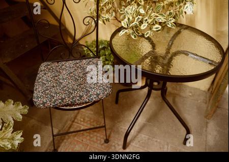 Metallstuhl mit Blumenpolster und Glastisch in einer gemütlichen Gartenecke. Konzept der Entspannung im Freien, Vintage-Möbel und ruhiger Innenhof Stockfoto
