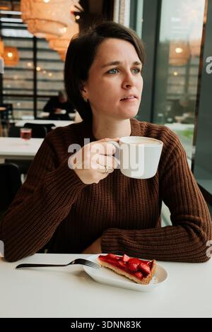 Eine Frau hält eine Tasse Cappuccino in der Nähe eines Fensters mit einer Erdbeertorte auf dem Tisch in einem modernen Café. Stockfoto