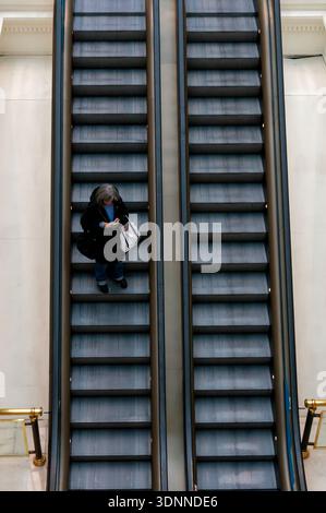 Eine Frau, die ein Smartphone benutzt, während sie eine symmetrische Rolltreppe in einem modernen Gebäude hinunterfährt. Draufsicht mit geometrischen Linien und Stadtverkehr Stockfoto