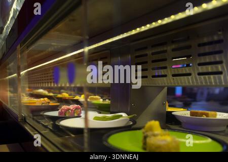 Eine perspektivische Nahansicht farbenfroher Platten mit verschiedenen Arten von Sushi, Rollen und Edamame, die sich auf einem rotierenden Förderband bewegen. Die Szene-Featur Stockfoto