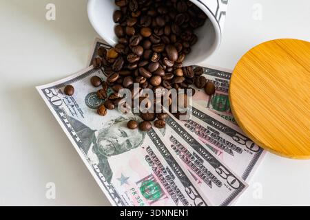 Ein hoch-winkliger konzeptioneller Schuss dunkel gerösteter Kaffeebohnen, die aus einem Keramikbehälter auf mehrere 50-Dollar-Banknoten überlaufen. Ein Bambusdeckel sitzt Stockfoto