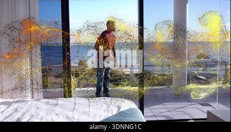 Stehende Frau in rotem Oberteil und Jeans, die Gerät auf Balkon, Bett, Glastüren, goldene Flecken hält Stockfoto