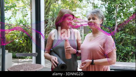Matte und Flasche halten, zwei Frauen im grauen Tank, rosa Oberteil plaudern auf der Terrasse, magentafarbene Überzüge Stockfoto
