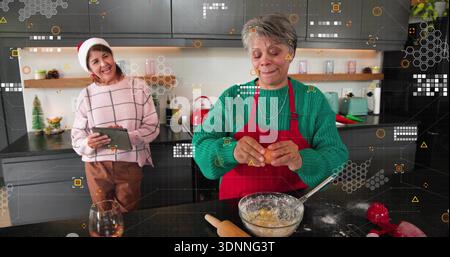 Knackende Seniorin in roter Schürze, grüner Pullover, der Ei in die Glasschale in der Küche mit Tablette gibt Stockfoto