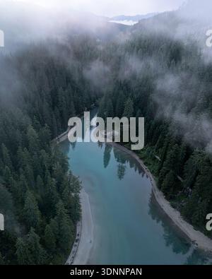 Aus der Vogelperspektive auf das türkisfarbene Wasser des Landrosees, das sich durch dichte Wälder schlängelt, umgeben von ätherischem Nebel und Bergen, Landro, Trentino-Südtirol, Stockfoto