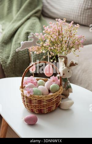 Osterkorb mit Ester-Eiern, Plüschhase und Frühlingsdekor auf weißem Tisch im Cozy Wohnzimmer Stockfoto