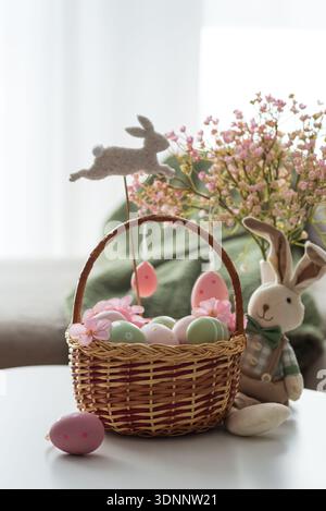 Osterkorb mit Ester-Eiern, Plüschhase und Frühlingsdekor auf weißem Tisch im Cozy Wohnzimmer Stockfoto