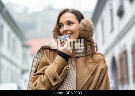 Eine Frau steht auf einer Stadtstraße und nimmt einen Happen von einem Dessert. Sie trägt einen warmen Mantel und Ohrenschützer, während sie lächelt. Gebäude umgeben sie und deuten auf ein ch hin Stockfoto