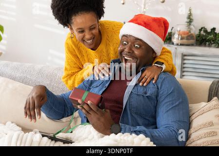 Afroamerikanisches Paar sitzt auf einem Wohnzimmer Sofa, Mann mit eingewickeltem Geschenk, trägt Weihnachtsmann-Hut Stockfoto