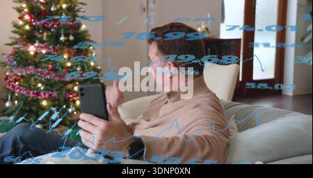 Das Smartphone hält, Mann in beigem Hemd, der auf dem Sofa Gesten mit Weihnachtsbaum und blauen Karten überlagert Stockfoto