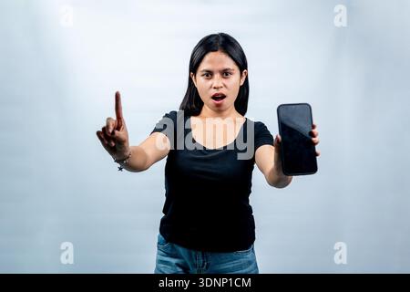 Porträt einer überraschten indonesischen Frau, die ein Mobiltelefon mit leerem Bildschirm hält und im Studio als Warnung oder Ratschlag mit dem Finger nach oben zeigt. Stockfoto