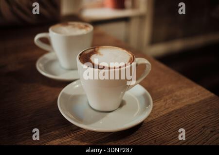 Zwei Tassen Cappuccino stehen auf einem Holztisch in einem gemütlichen Café mit sanftem Licht. Konzept von warmer Atmosphäre, Kaffeekultur, langsamen Momenten und Stockfoto