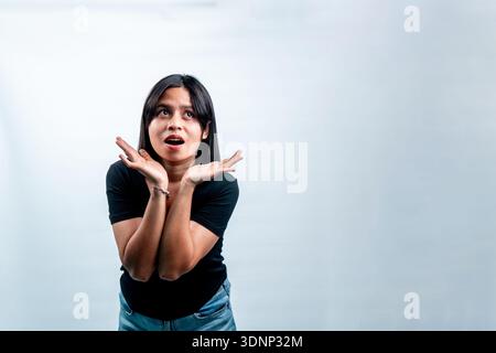 Überraschte indonesische Frau in einem schwarzen T-Shirt, die mit offenem Mund aufblickt und staunende Hände, isoliert auf weiß. Stockfoto