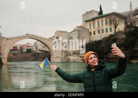 Eine Frau macht ein Selfie mit der Flagge Bosnien und Herzegowinas in der Nähe der Alten Brücke am Fluss. Konzept des modernen Reisegeschichtenerzählens, soziale Medien Stockfoto