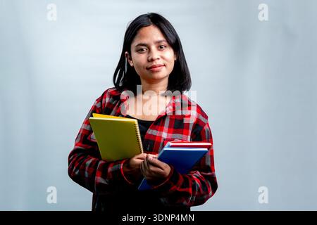 Porträt einer jungen südostasiatischen Frau in einem roten karierten Hemd, die steht und bunte Notizbücher hält, während sie in die Kamera schaut Stockfoto