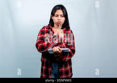 Eine nachdenkliche junge südostasiatische Frau in einem roten karierten Hemd, die einen Stift auf ihr Kinn legt und ein Notizbuch vor einem schlichten Hintergrund hält. Stockfoto