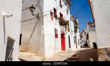 Charmante, enge Straße in Altea, Alicante, mit weiß getünchten Gebäuden, knallroten Türen und lebendigen Blumen. Typisch spanische Architektur unter einem Klo Stockfoto