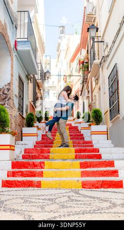 Ein Paar teilt einen romantischen Moment auf den farbenfrohen Stufen, die im Muster der spanischen Flagge gemalt sind. Das Hotel liegt in den malerischen Straßen von Alicante, Spanien, Stockfoto