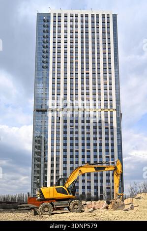 Ein großer Bagger arbeitet auf einer Baustelle und bereitet den Boden für eine neue Wohnbauentwicklung vor. Moderne Hochhausstruktur sichtbar Stockfoto