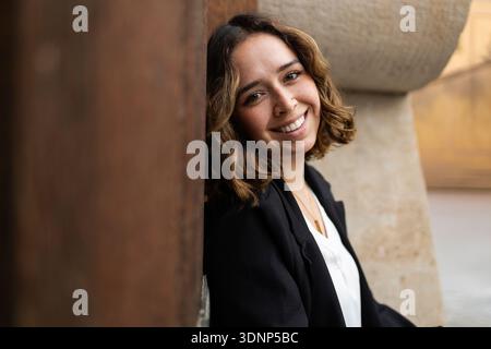 Eine junge Frau mit welligen Haaren lächelt warm, während sie sich draußen an eine rustikale braune Wand lehnt. Sie trägt einen schwarzen Blazer über einem weißen Oberteil und strahlt ein cas aus Stockfoto