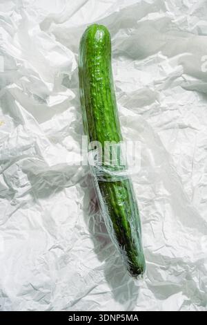 Eine frische Gurke, in Kunststoff gewickelt, liegt elegant auf zerknittertem weißem Papier. Der Kontrast zwischen dem leuchtenden Grün und dem neutralen Hintergrund Stockfoto
